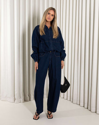 Person wearing a blue denim outfit with high-waisted jeans and a jacket against a white curtain background.