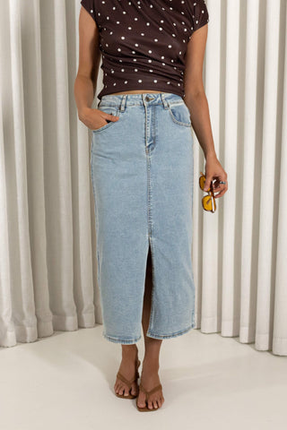 A model wearing a light-wash blue denim midi skirt with a front slit, paired with a brown polka-dot top and brown sandals, standing in front of white pleated curtains.