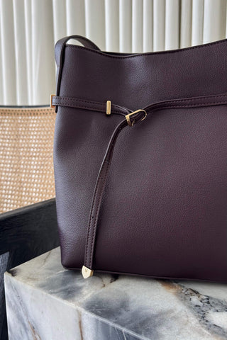 A dark plum purple pebbled leather tote bag with a front strap detail and gold-tone hardware, resting on a marble surface.