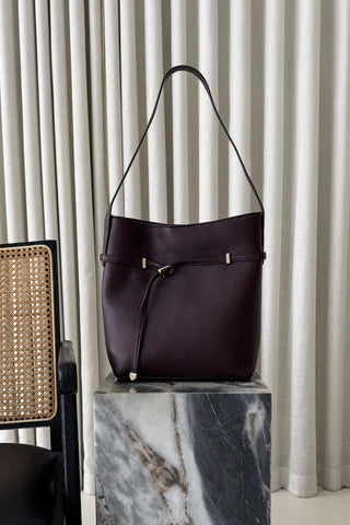 A dark plum leather bucket bag with a drawstring detail and small gold-tone hardware, displayed on a marble block pedestal in front of beige curtains.