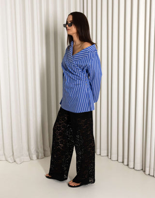 Woman wearing a blue striped shirt and black lace pants against a white curtain background