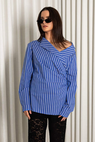 A woman modeling a blue and white vertically striped asymmetrical wrap top with long sleeves, paired with black lace pants and black sunglasses, standing in front of a white pleated curtain.