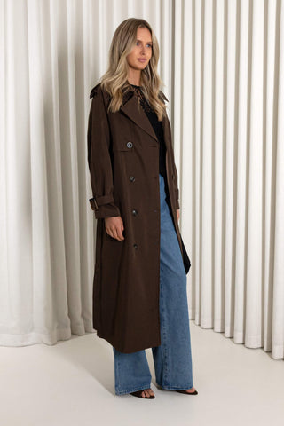 A woman modeling a long, double-breasted brown trench coat over a black top and wide-leg blue jeans, standing in front of a white curtain backdrop.
