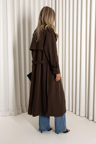 A woman shown from the back wearing a long dark brown trench coat with a matching belt, blue jeans, and brown heeled sandals, standing in front of a white pleated curtain backdrop.