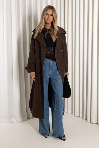 A woman modeling a long brown trench coat over a black lace top and wide-leg blue jeans, standing against a white pleated curtain backdrop.
