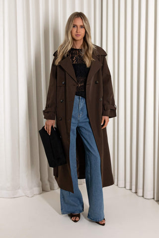 A woman models a long brown double-breasted trench coat over a black lace top and wide-leg blue jeans, holding a black clutch bag against a background of white vertical drapes.