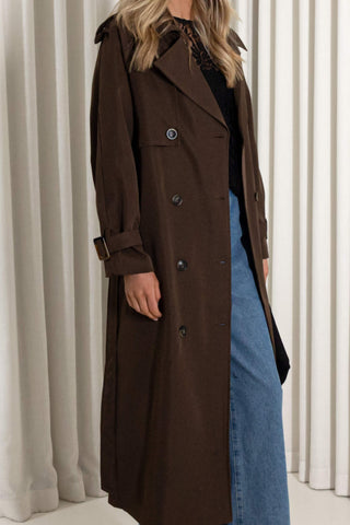 A woman wearing a long dark brown double-breasted trench coat over a black top and blue denim skirt, standing in front of white pleated curtains.