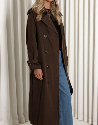 Person wearing a brown trench coat over a black top and blue jeans against a white curtain background