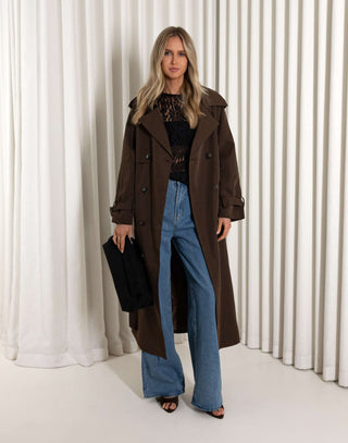 Woman wearing a brown coat and blue jeans standing against a white curtain background