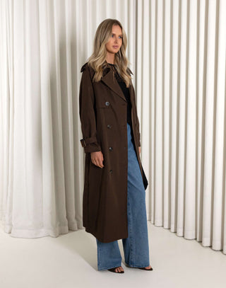 Woman wearing a long brown coat and blue jeans standing against a white curtain.