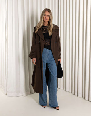 Woman wearing a brown coat and blue jeans standing against a white curtain.