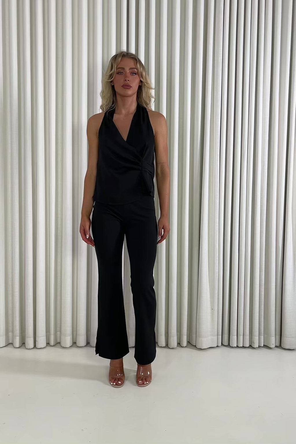 Woman wearing a black jumpsuit standing against a white curtain background Andy black V-neck top and Cole black high-waist tailored flare pants