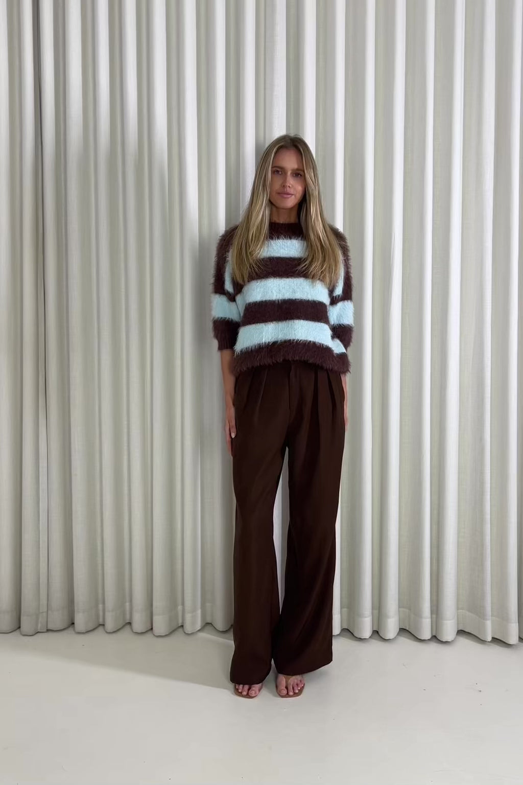 Woman wearing a brown and blue striped sweater with wide-leg pants against a white curtain background
