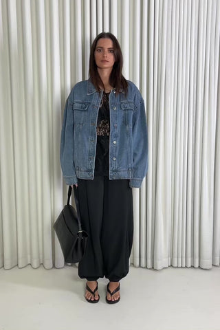 Woman wearing a denim jacket and black pants standing against a white curtain background