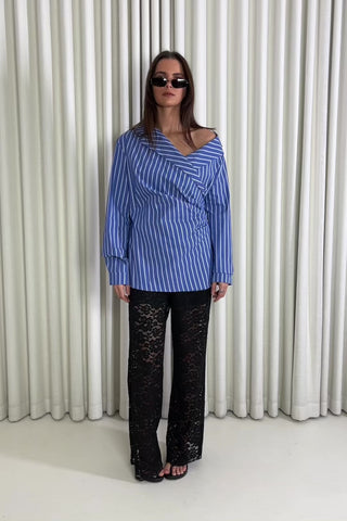 Woman wearing a blue striped shirt and black lace pants against a white curtain background
