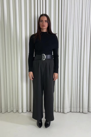 Woman wearing a black turtleneck and black pants with a belt against a white curtain background