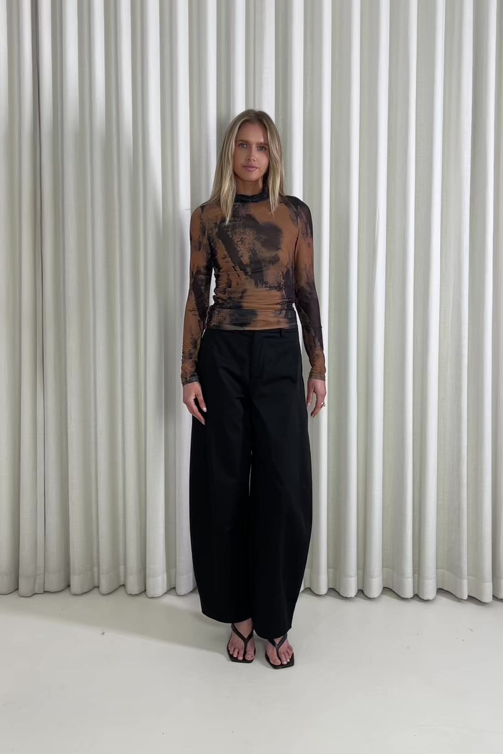 Woman wearing a black and brown tie-dye top with high-waisted black pants against a white curtain background.