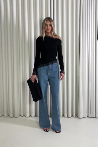 Woman wearing a black top and blue jeans standing against a white curtain, showing of the outfit.