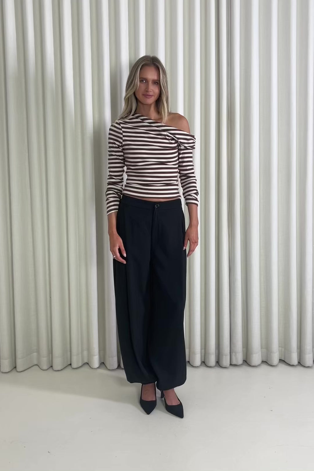Woman wearing a striped top and black pants standing against a white curtain.