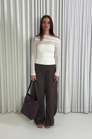 Woman wearing a cream top and brown pants standing against a white curtain.
