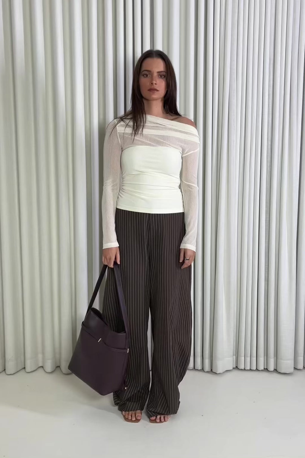 Woman wearing a cream top and brown pants standing against a white curtain.