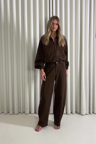 Woman wearing a brown jacket and jeans standing against a white curtain, showing of the outfit.