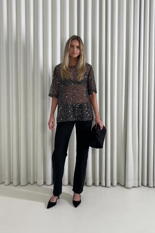 Woman wearing a sequin top and black pants against a white curtain background
