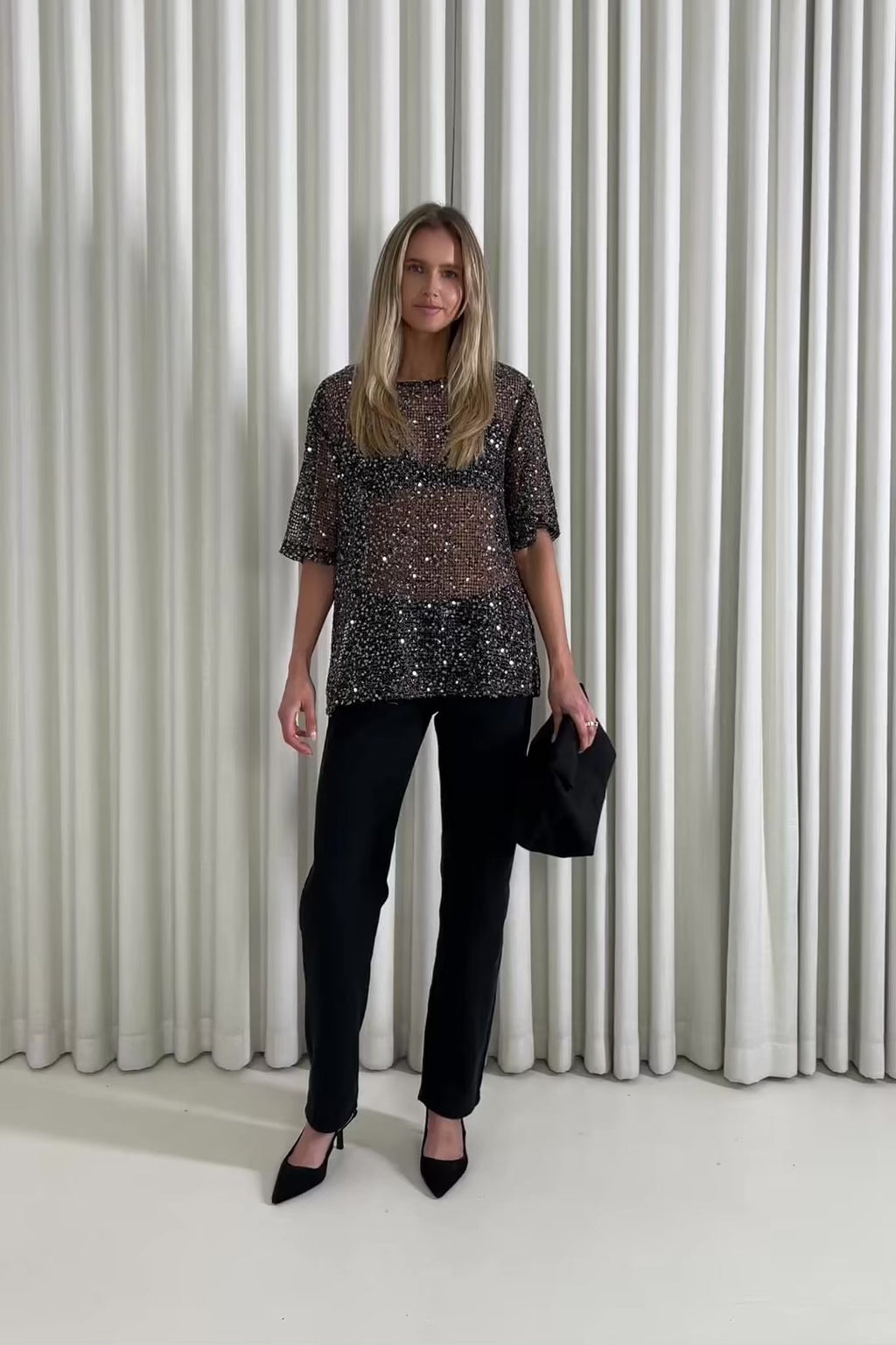 Woman wearing a sequin top and black pants against a white curtain background