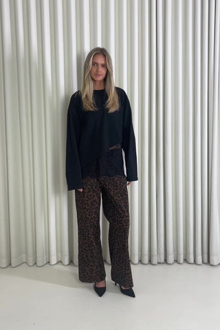 Woman wearing a black lace top and leopard print pants standing against a white curtain.