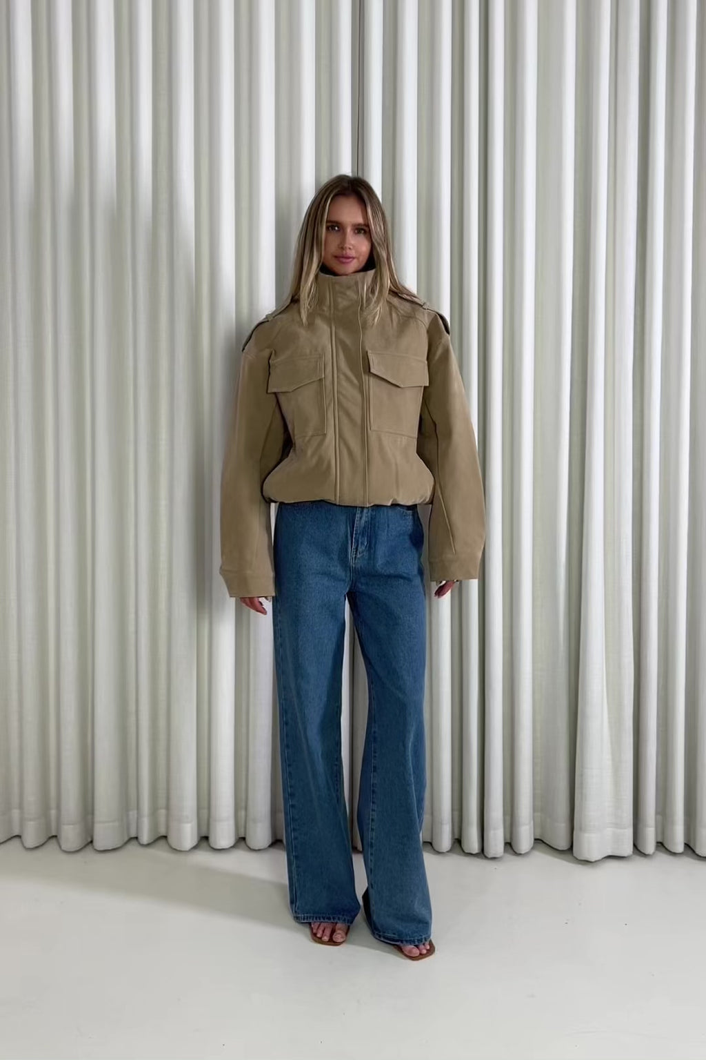 Person wearing a beige bomber jacket and blue jeans standing in front of white curtains