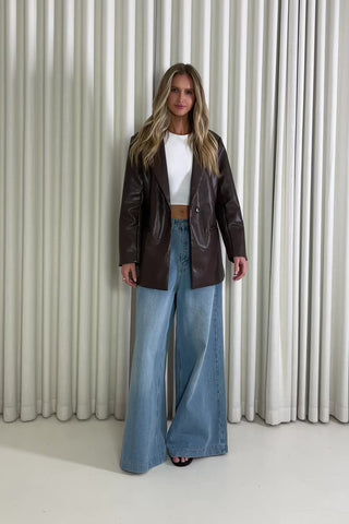 Woman wearing a brown leather jacket and blue jeans against a white curtain background