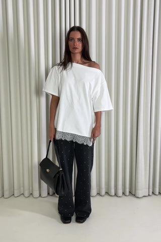 Woman wearing a white off-shoulder top and black pants with a lace trim, standing against a white curtain.