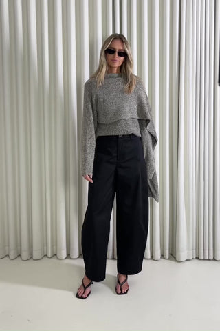 Woman wearing a knit and black pants standing against a white curtain, showing of the outfit.