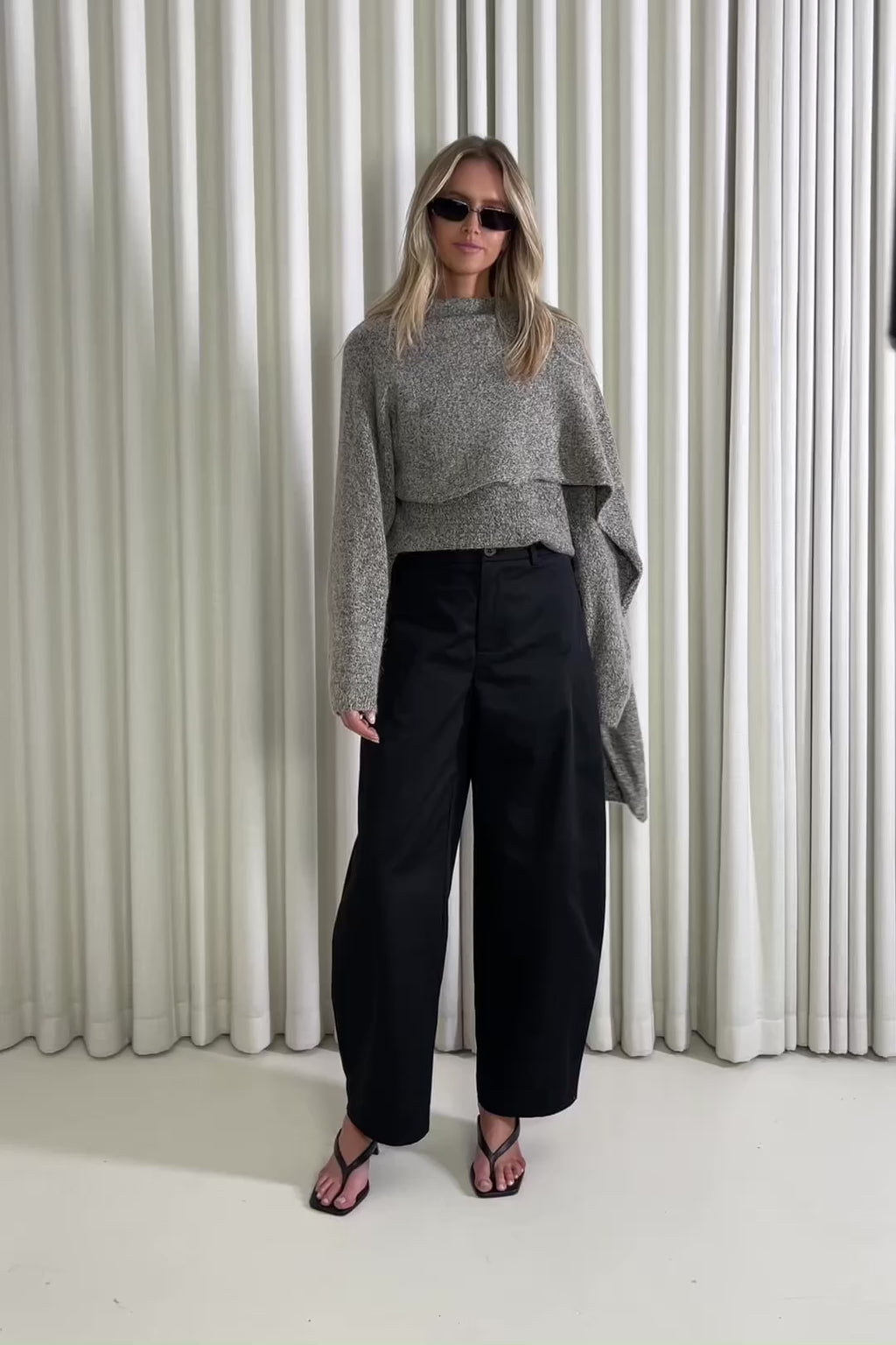 Woman wearing a knit and black pants standing against a white curtain, showing of the outfit.