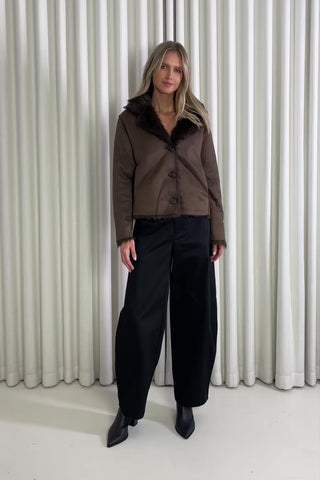Woman wearing a brown jacket and black pants standing against a white curtain, showing of the outfit.