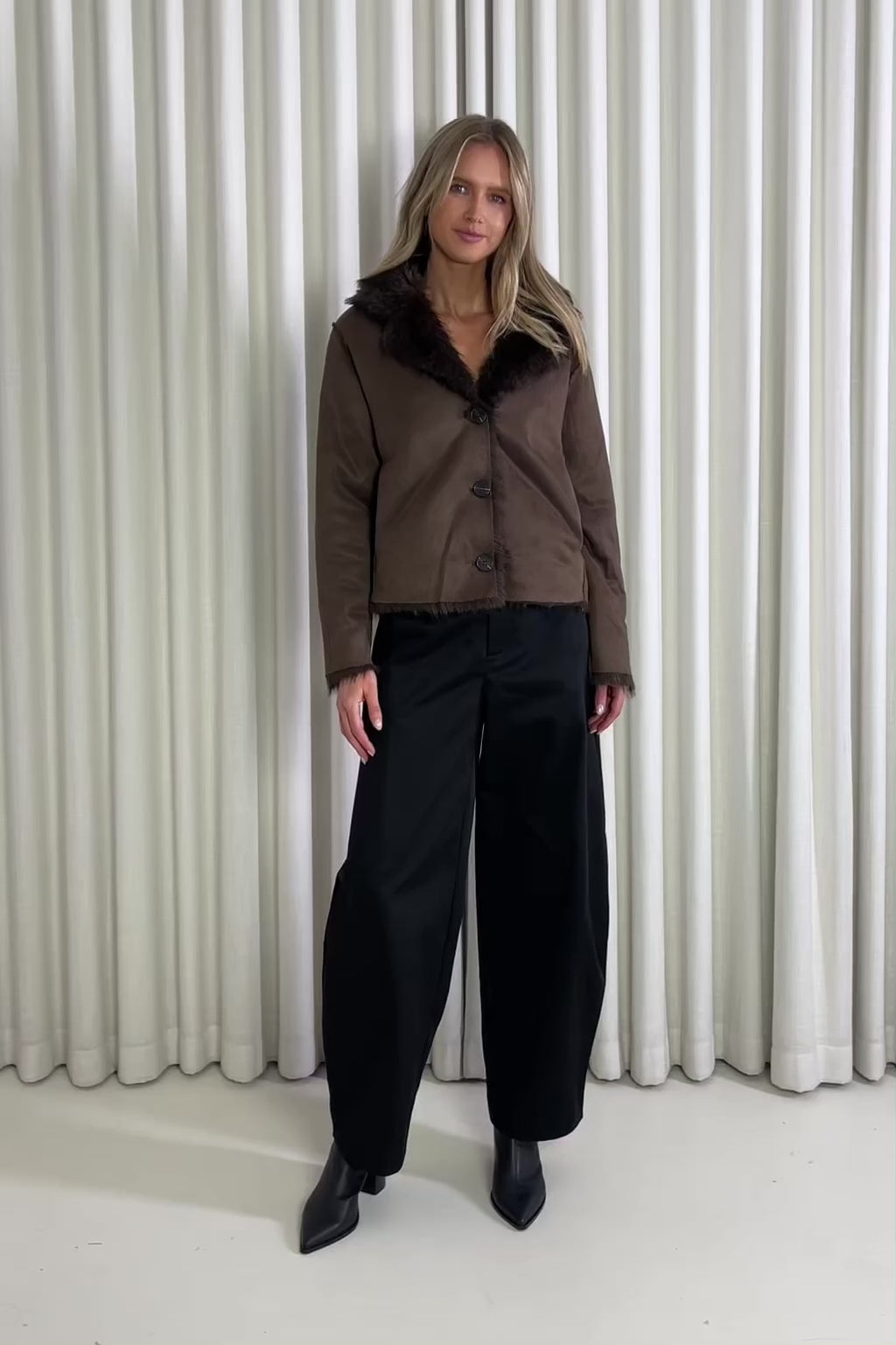 Woman wearing a brown jacket and black pants standing against a white curtain, showing of the outfit.