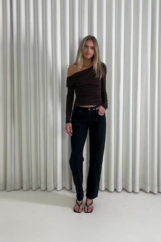 Woman wearing a brown off-shoulder top and black pants against a white curtain background