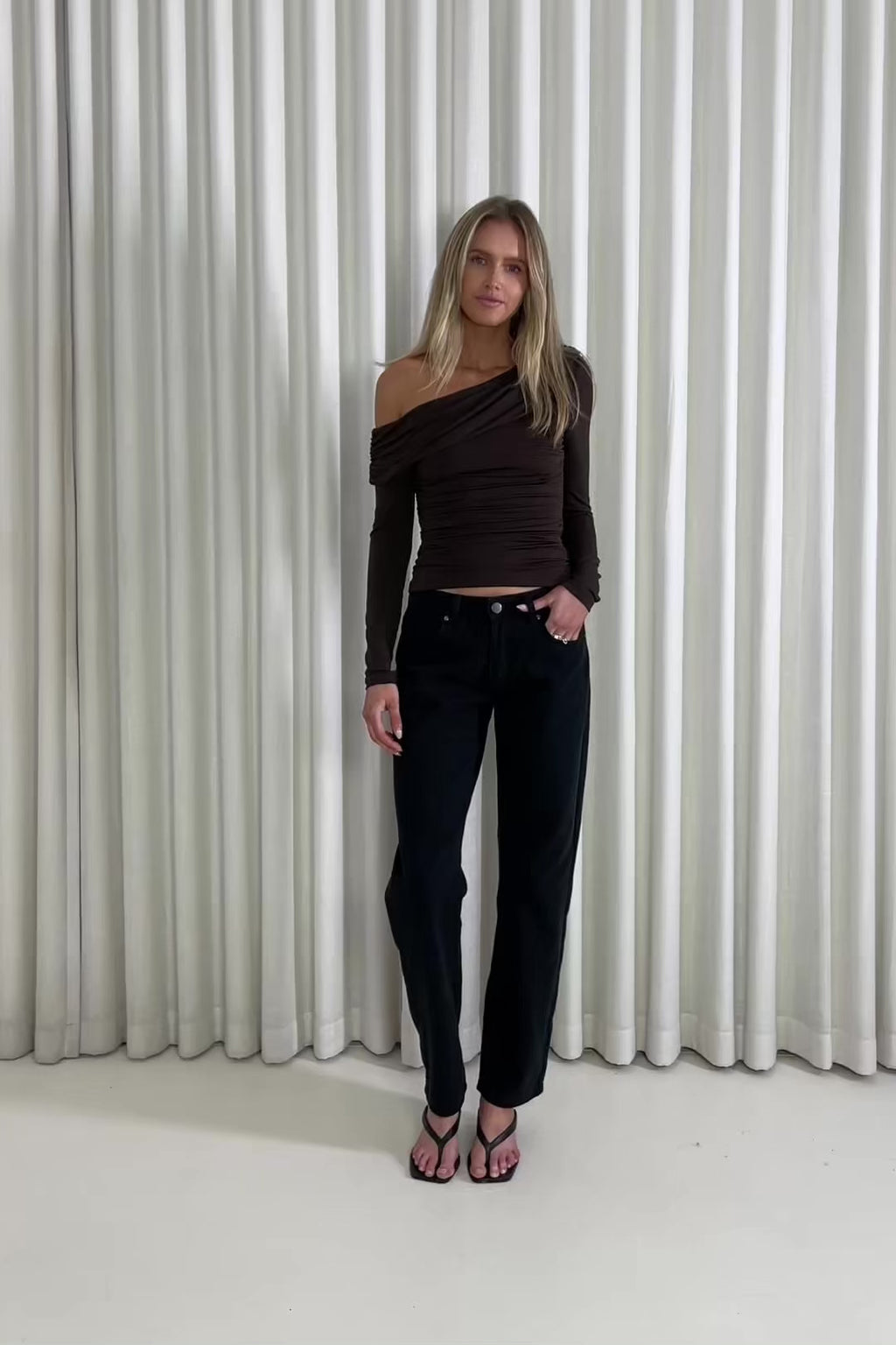 Woman wearing a brown off-shoulder top and black pants against a white curtain background