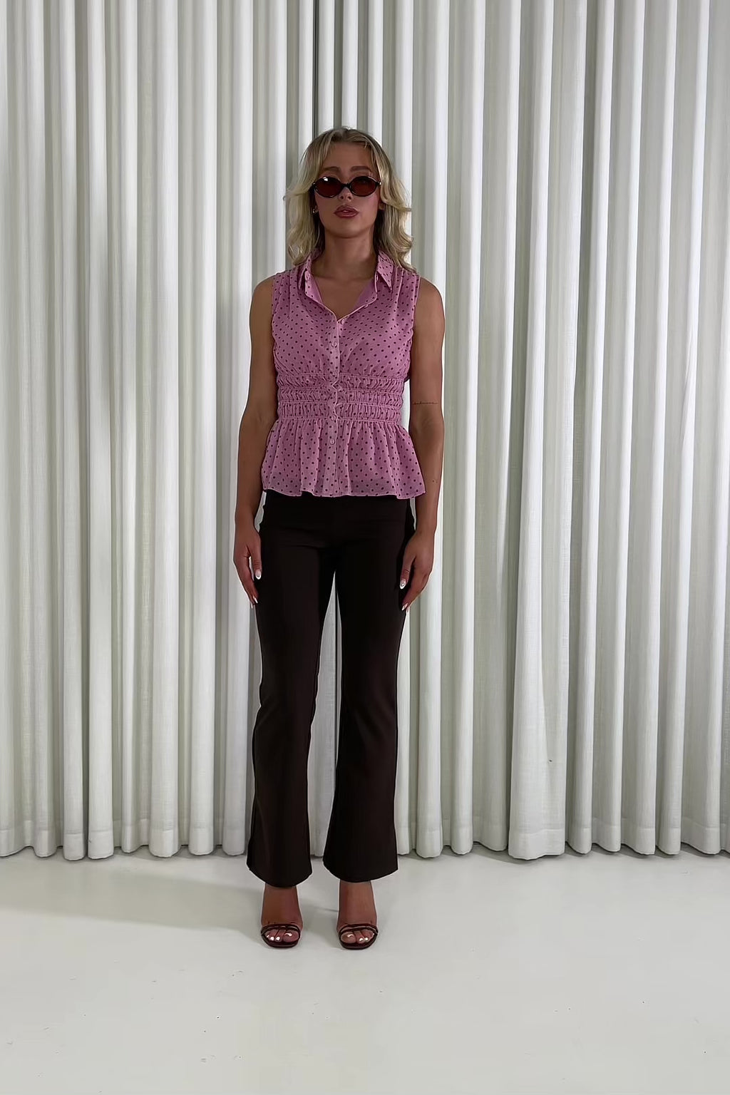 Woman wearing a pink sleeveless top and brown pants against a white curtain background Penny pink polka dot collared top with shirred waist Cole brown high-waist tailored flare pants with leg split