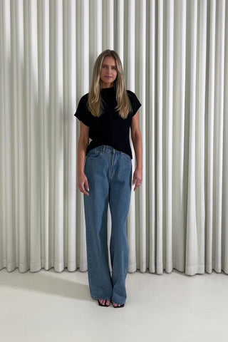 Woman wearing a black knit top and blue jeans standing against a white curtain background