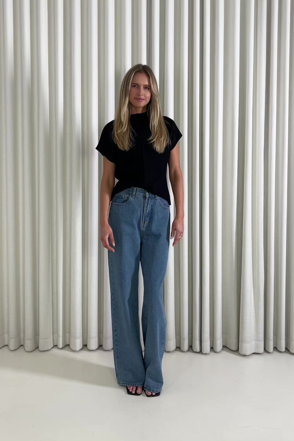 Woman wearing a black knit top and blue jeans standing against a white curtain background