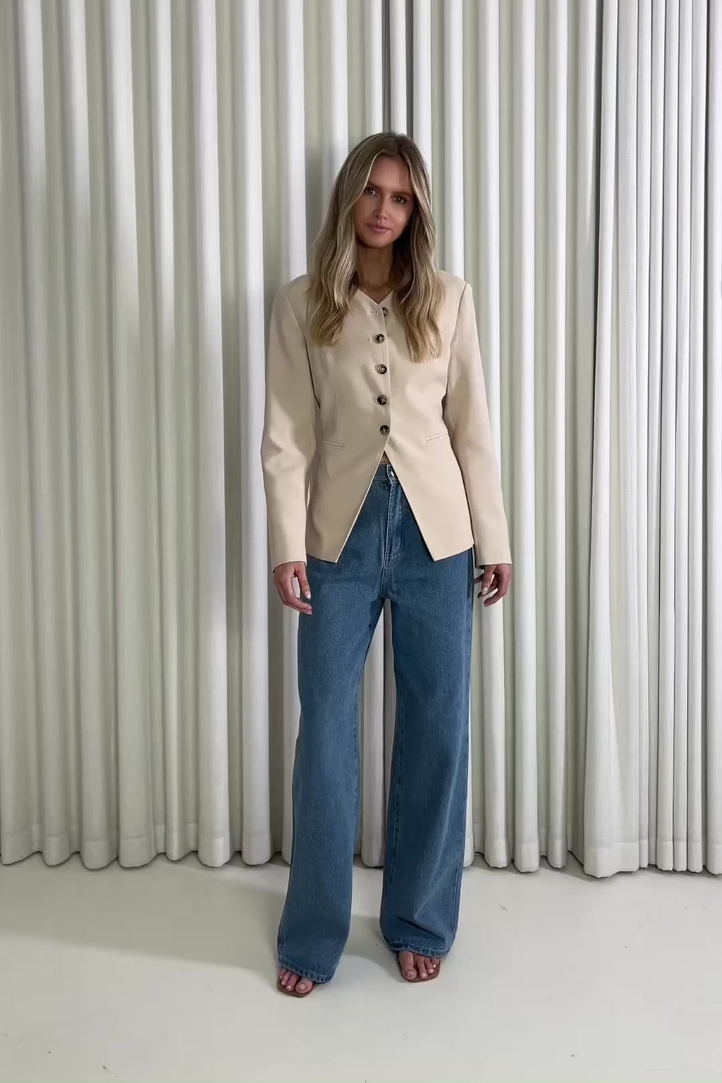 Woman wearing a beige blazer and blue jeans standing against a white curtain, showing of the outfit.