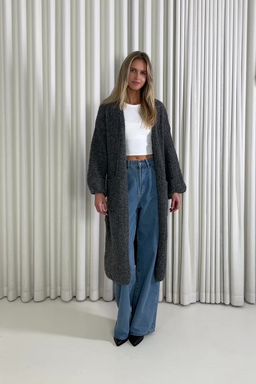 Woman wearing a long dark gray cardigan, white top, and blue jeans against a white curtain background.