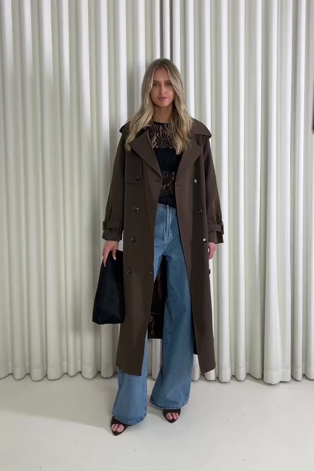 Person wearing a brown trench coat over a black top and blue jeans against a white curtain background