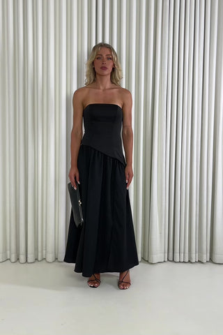 Woman wearing a black strapless dress with a slit, holding a black clutch against a white curtain background. Chris black dress with contrasting fabric and skirt split