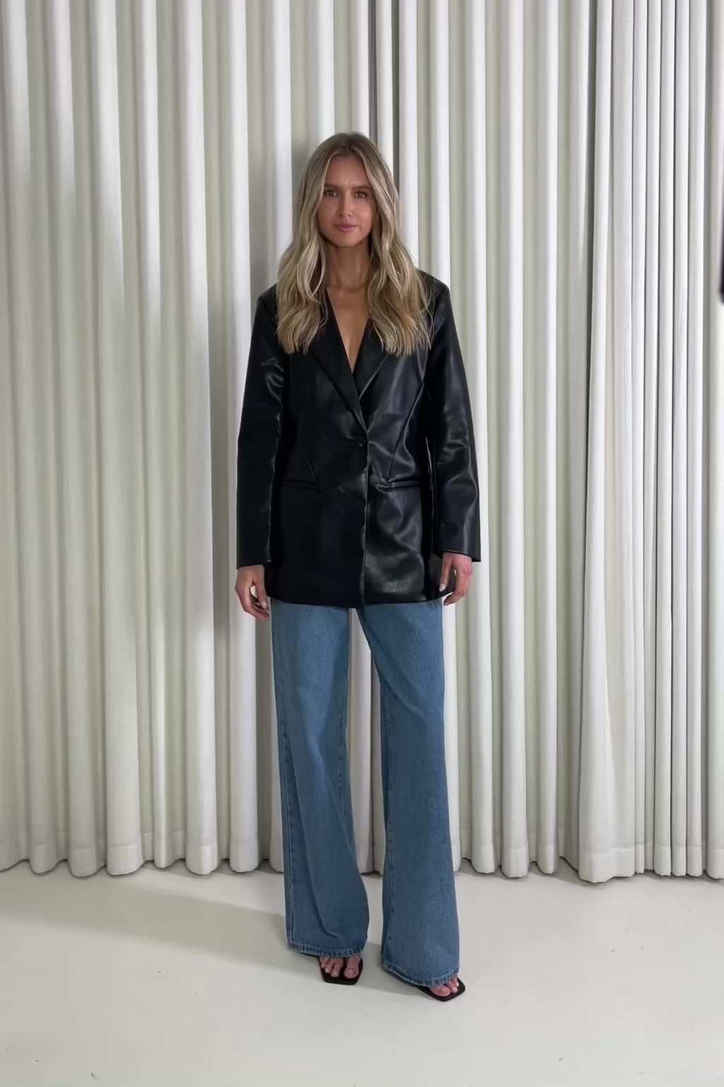 Woman wearing a black blazer and blue jeans standing against a white curtain, showing of the outfit.