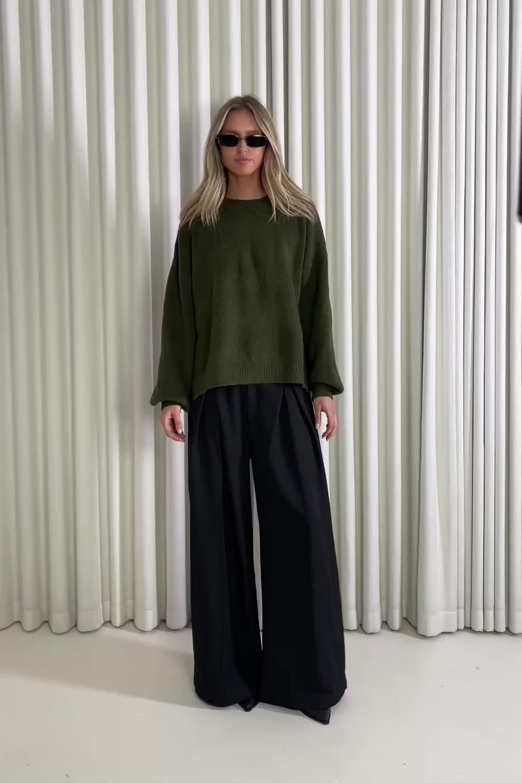 Woman wearing a green knit and black pants standing against a white curtain, showing of the outfit.