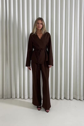 Woman wearing a brown blazer and brown matching pants standing against a white curtain background