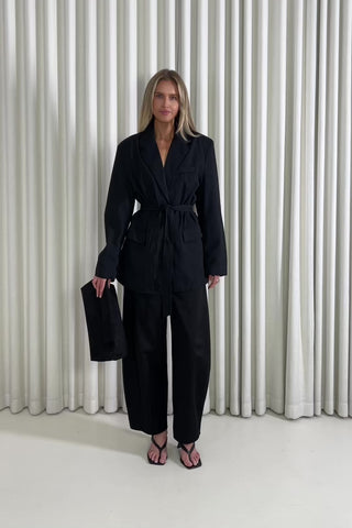 Woman wearing a black tie blazer and black pants standing against a white curtain background
