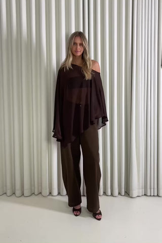 Woman wearing a sheer cape top and brown jeans standing against a white curtain, showing of the outfit.